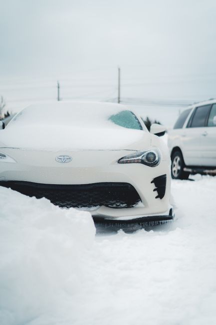 A white Toyota car covered with snow sits in a snowy outdoor parking area, with the front of the vehicle partially obscured by snow accumulation on the ground. In the background, another white vehicle is parked alongside, and utility poles with power lines stretch across a cloudy sky, suggesting cold winter weather. The snowy surface shows patterns of footprints and tire tracks, indicating recent activity. This scene captures the quiet and cold atmosphere of a winter day, emphasizing the need for outdoor maintenance and garden clearance as part of seasonal preparations. Gardeners Edgware recommends these tips to keep outdoor spaces tidy and well-maintained, even in winter conditions.